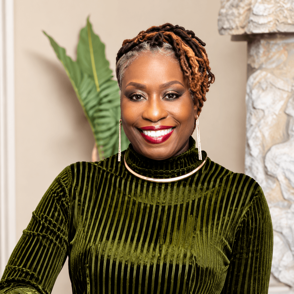 Smiling Black woman with styled locs, red lipstick, and a green velvet turtleneck.
