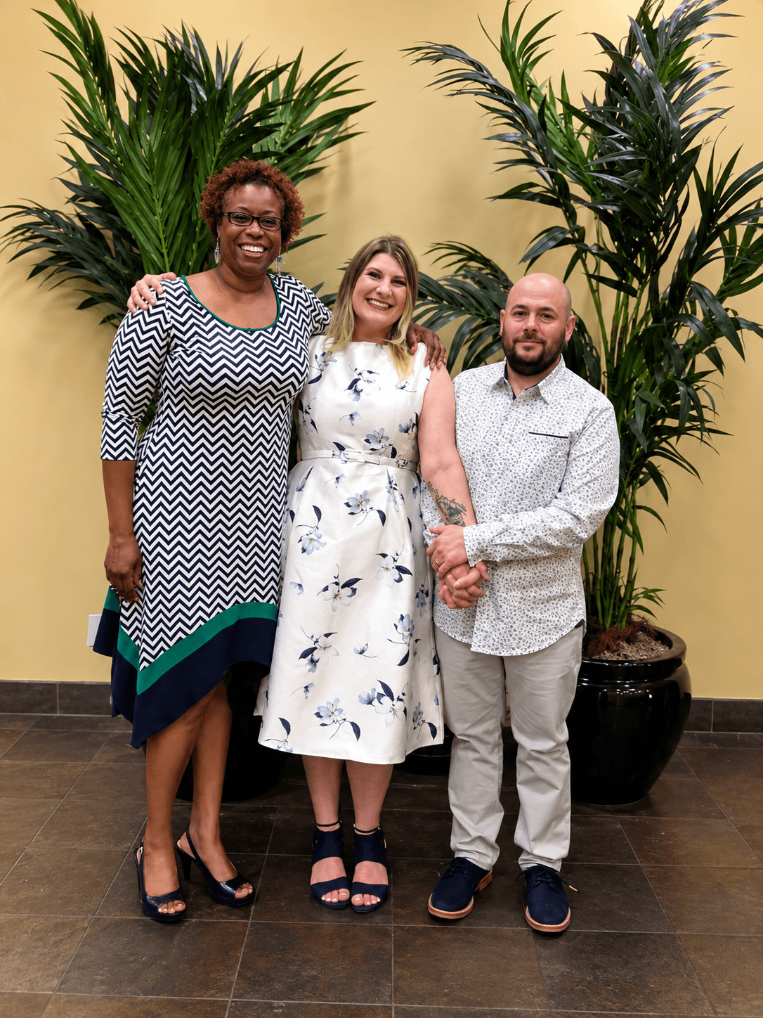 Three smiling people in patterned clothing pose together between two large potted palm plants.