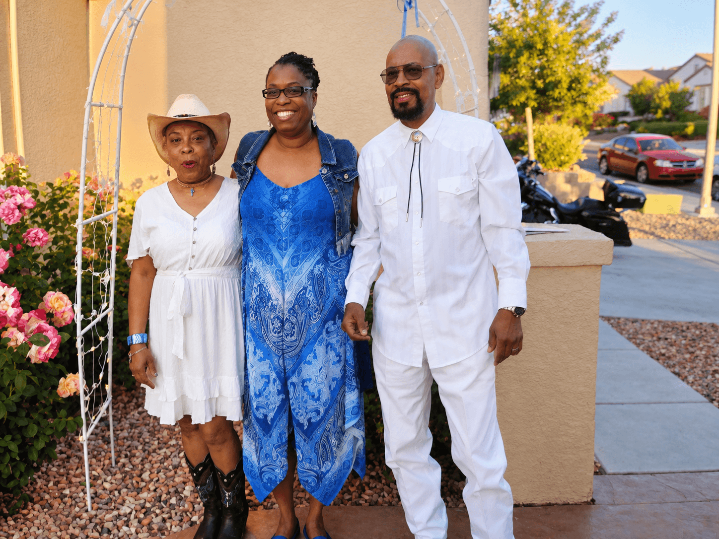 Three Black people pose outdoors. One wears a cowboy hat, another a blue patterned dress.